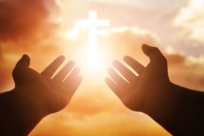 Silhouette hands with cross against cloudy sky during sunset