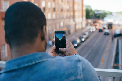 Rear view of man photographing city street from mobile phone while standing on footbridge