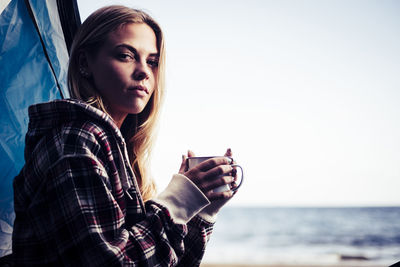 Young woman holding coffee cup by tent at beach against clear sky