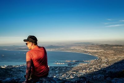 Rear view of young man sitting on cliff by sea against blue sky