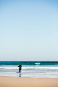 Rear view of man with surfboard walking at beach against clear sky