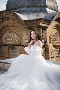Bride in a white dress sits on a ledge with an old building in the background. young caucasian woman