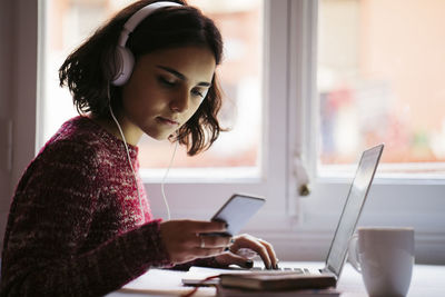 Young woman with laptop and headphones looking at cell phone