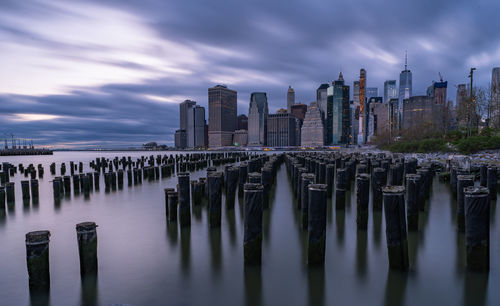 Wooden posts in river by buildings against sky at dusk