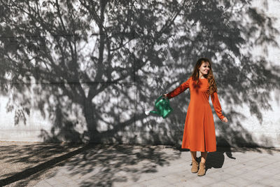 Young woman watering tree shadow while standing against wall