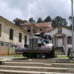 Mother and son sitting on vintage car outside building against sky