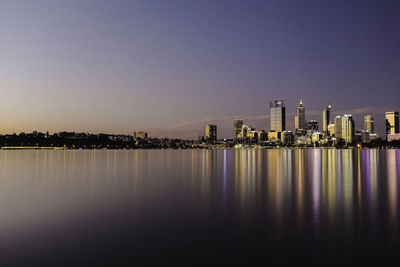 Illuminated buildings by river against clear sky at sunset