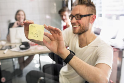 Young businessman sticking adhesive note on transparent glass with colleagues at background in new office