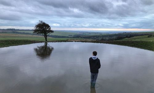 Rear view of teenage boy standing in lake against cloudy sky