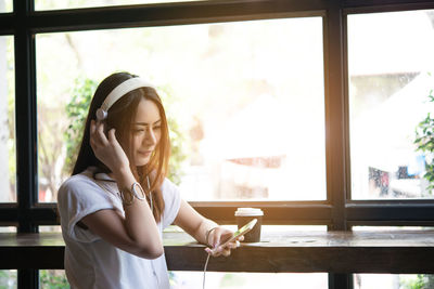 Young woman listening music through headphones by window