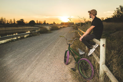 Side view of man sitting with bicycle on road during sunset