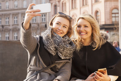 Smiling young woman taking selfie with friend from smart phone while sitting in city