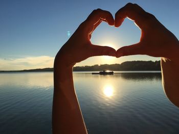 Cropped hand of person making heart shape against lake and sky during sunset