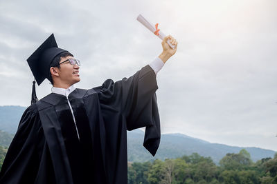 Happy young man in graduation gown standing on mountain against cloudy sky