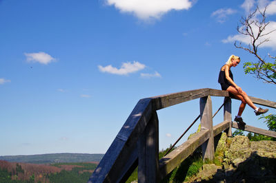 Low angle view of young woman sitting on railing against blue sky during sunny day