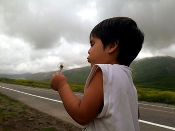 Boy blowing dandelion against cloudy sky
