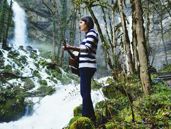 Young woman playing guitar while standing against waterfall in forest