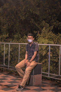 Portrait of young man sitting on railing against trees