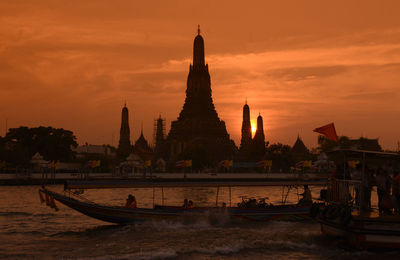 Silhouette man sailing boat in river with temple in background against orange sky