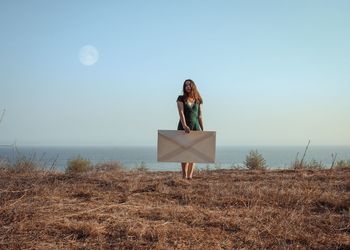 Woman holding large envelope while standing on field against sea and clear sky