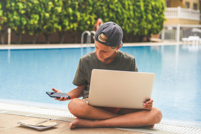 Boy using laptop while sitting by swimming pool