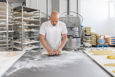 Focused bald male baker rolling dough while preparing baguette on metal table with tray during work in professional light bakehouse