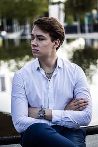 Thoughtful young man with arms crossed sitting against lake in park