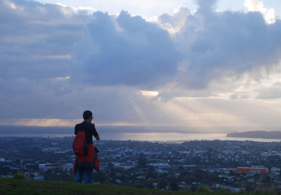 Rear view of man on mountain looking at cityscape against cloudy sky