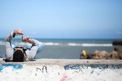 Mature man using smart phone while lying on retaining wall at beach against blue sky during sunny day