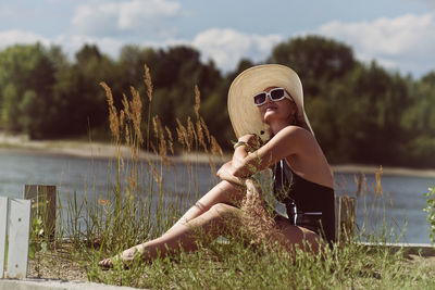 A woman in a swimsuit, hat and sunglasses sunbathes in summer on the riverbank among the grass