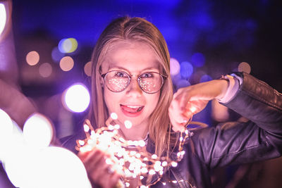 Portrait of young woman holding illuminated string light at night