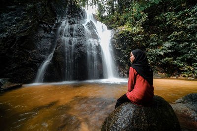 Rear view of girl sitting against waterfall in forest