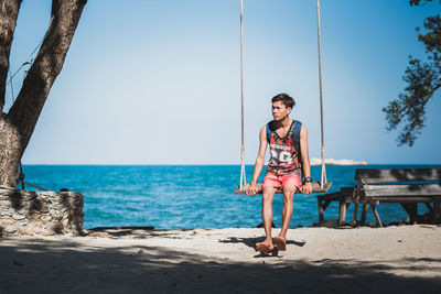 Full length of man sitting on swing at sea shore against sky