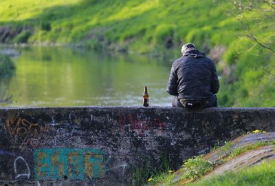 Rear view of man sitting on retaining wall by river