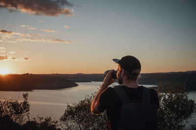 Rear view of man standing on shore against sky during sunset