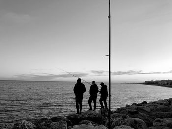 Rear view of men fishing while standing on rocks by sea