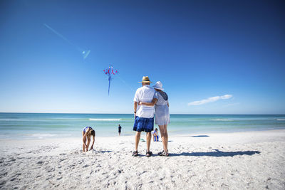 Grandparents standing while grandchildren playing at beach against sky during sunny day