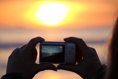 Cropped image of woman hand photographing sea against sky during sunset