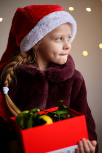 A brooding christmas girl in santa's red cap holds a red box of tangerines.