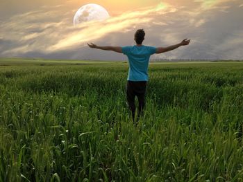 Rear view of man standing with arms outstretched on field against moon in sky