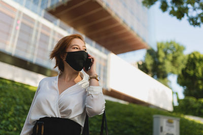 Low angle of determined female executive manager wearing medical mask walking while speaking on the mobile phone on street and looking away