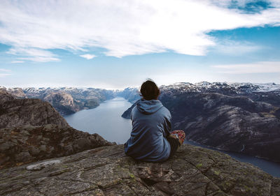 Rear view of woman sitting on rock while looking at fjord amidst rocky mountains against sky