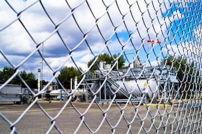 Industry seen through chainlink fence against cloudy sky