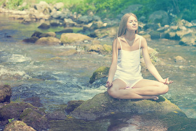 Young woman meditating while sitting on rock by river