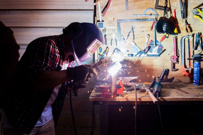 Side view of mature male worker using angle grinder while working with metal detail with glowing sparks in garage