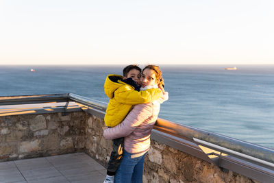 Woman with child in arms standing on rooftop with the sea in background