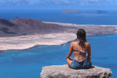 Rear view of young woman overlooking sea while sitting on rock at lanzarote