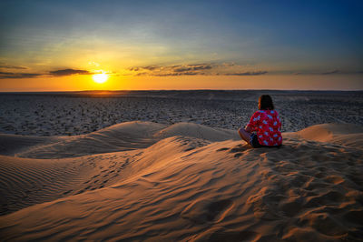 Rear view of woman sitting on sand at beach against sky during sunset