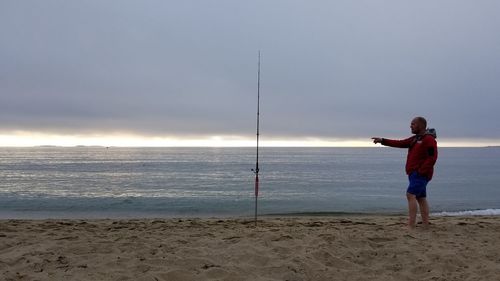 Full length of man pointing at fishing rod in sand against cloudy sky during sunset
