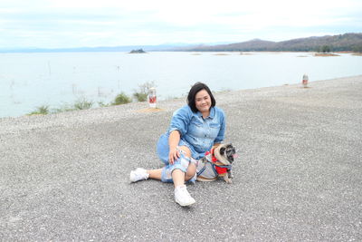 Portrait of woman sitting with pug by sea on road 